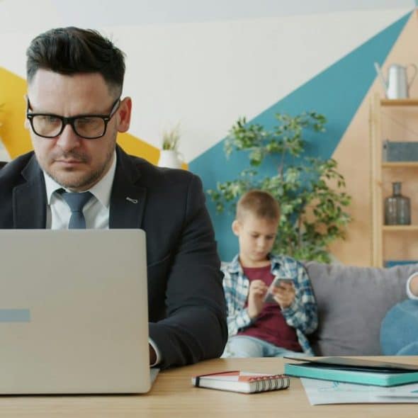 A man works on a laptop while a woman reads and a boy uses a phone behind him.
