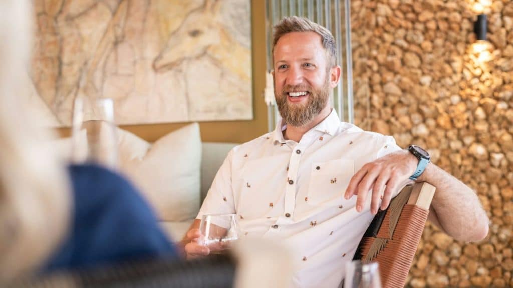 A smiling, bearded man in a patterned white shirt holds a glass while sitting.