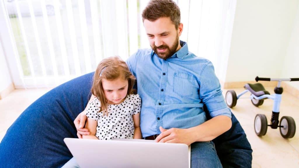 A father and young daughter sit together on a blue beanbag chair, looking at a laptop.