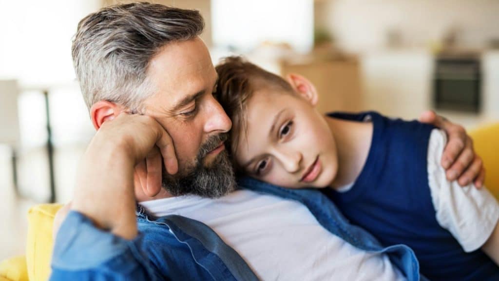 Middle-aged man with a beard and graying hair sitting with his son leaning on his shoulder.