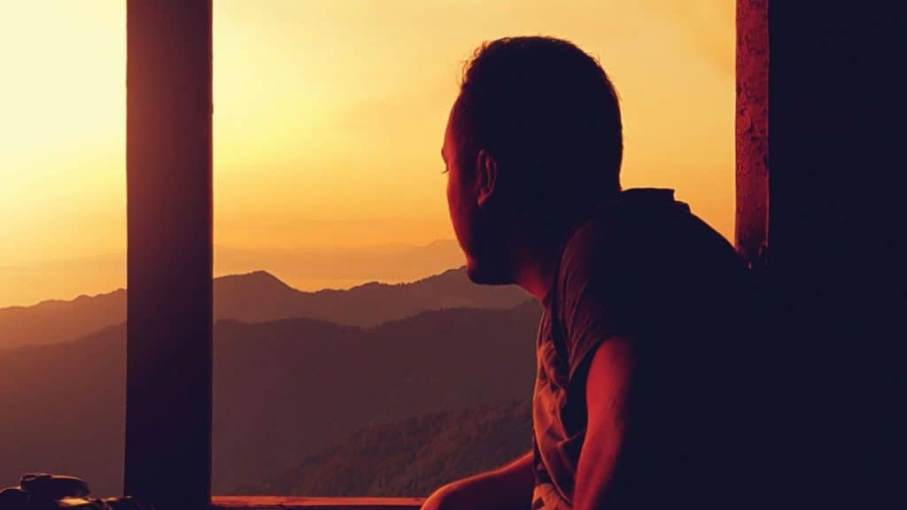 A silhouetted man looks out a window at a mountain range during sunset.