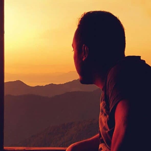 A silhouetted man looks out a window at a mountain range during sunset.