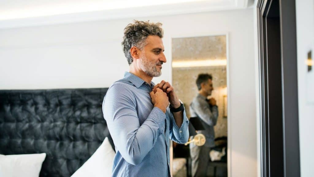 Middle-aged man with gray hair buttoning his blue shirt in front of a mirror.