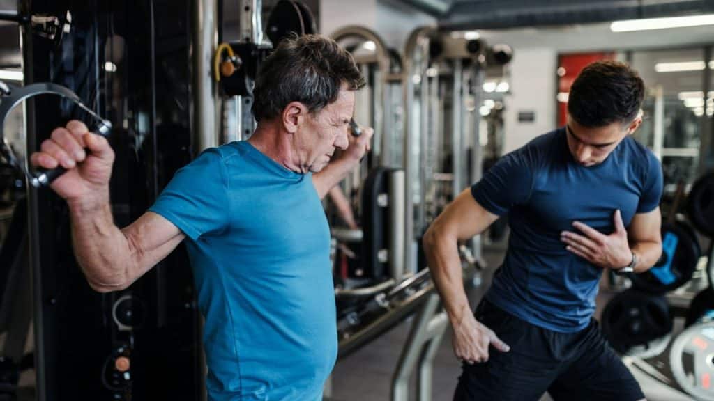 A young personal trainer is instructing an older man on a cable machine.