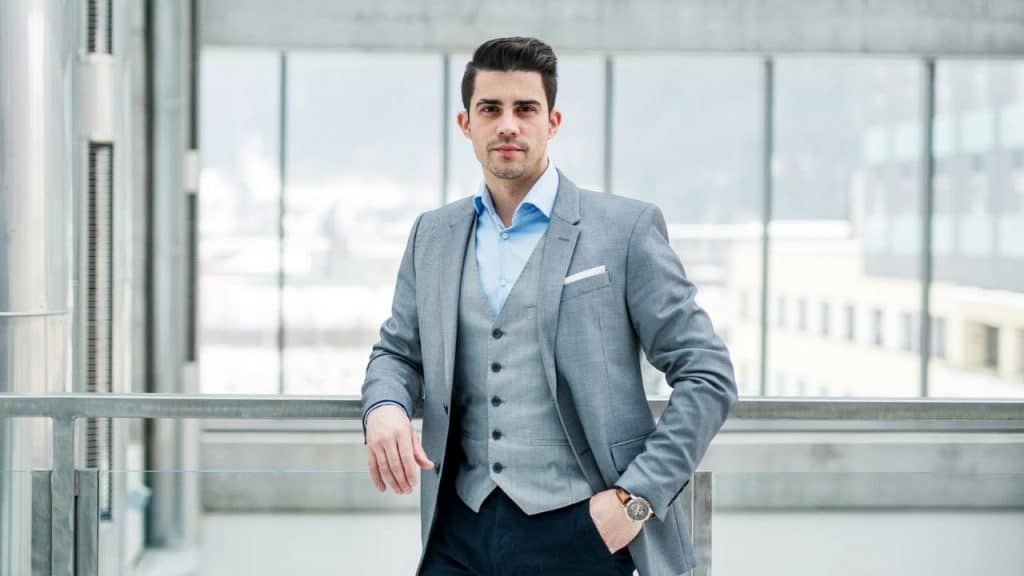 A handsome young businessman in a three-piece suit is standing confidently indoors.