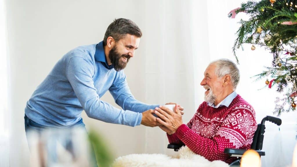 Younger man hands a cup to an older man in a wheelchair, smiling by a Christmas tree.