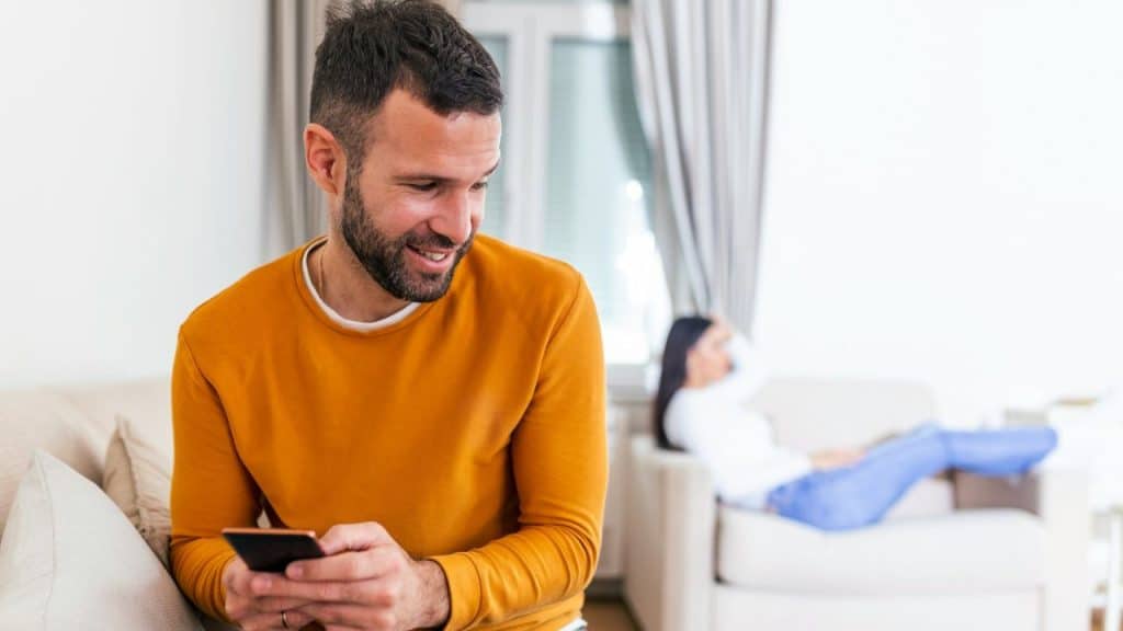 A man in an orange shirt smiles while looking at his smartphone on a couch.