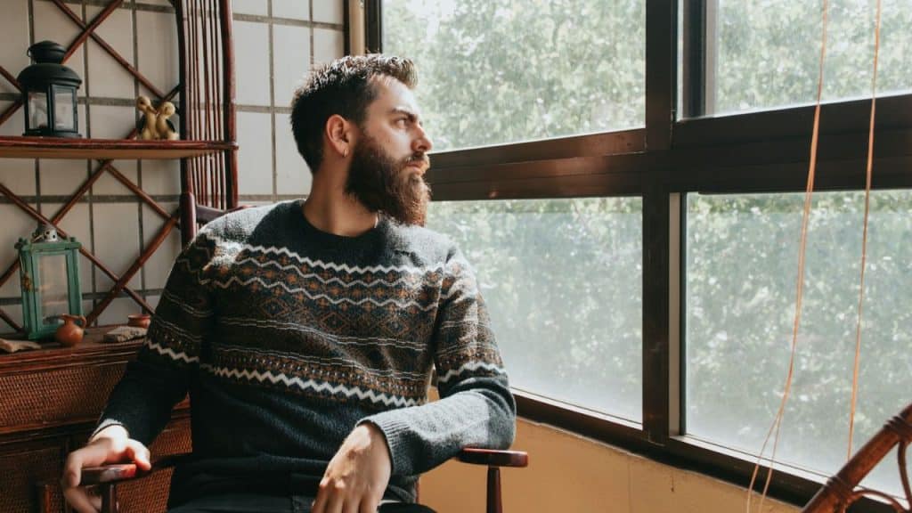 Bearded man in a patterned sweater sitting by a large window looking out at trees.