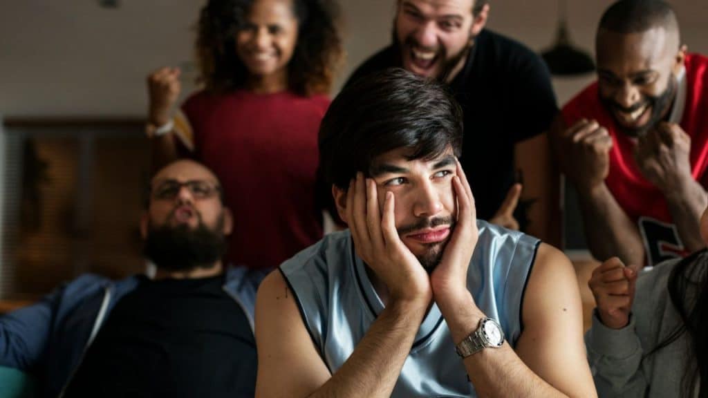 A man sits dejected with his hands on his face while five cheering people stand behind him.