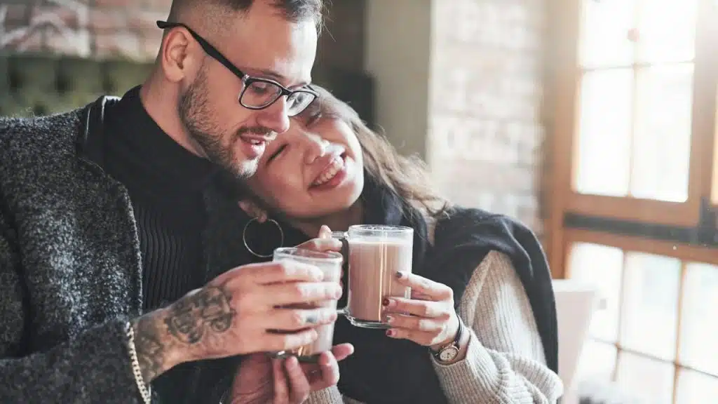 A young, tattooed couple smiles while holding and clinking glasses of hot cocoa indoors.