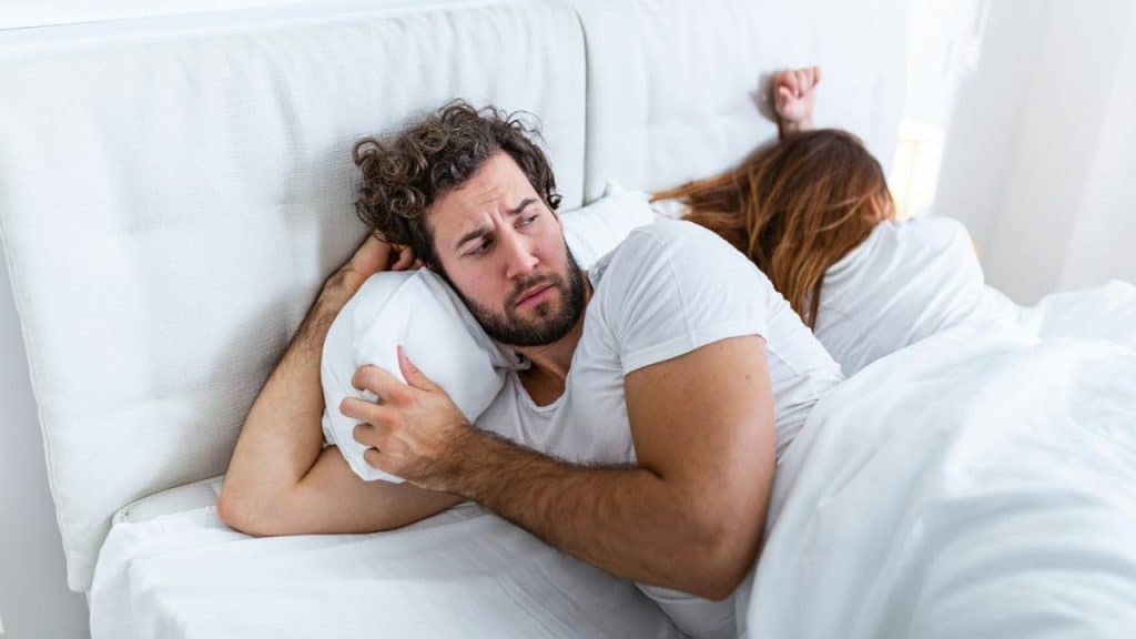 A man sits awake and pensive in an all-white bed with a woman facing away.