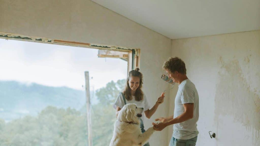 A couple smiling while painting a room with their dog beside them.