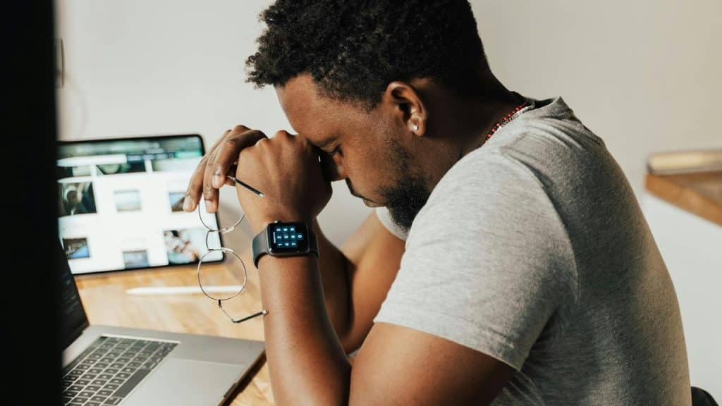 A man sitting at a desk with his head down holding his glasses looking tired.