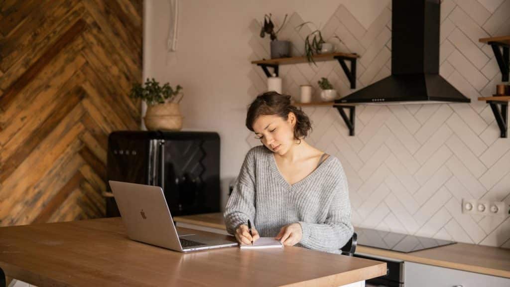 A woman sitting at a kitchen table writing in a notebook beside a laptop.