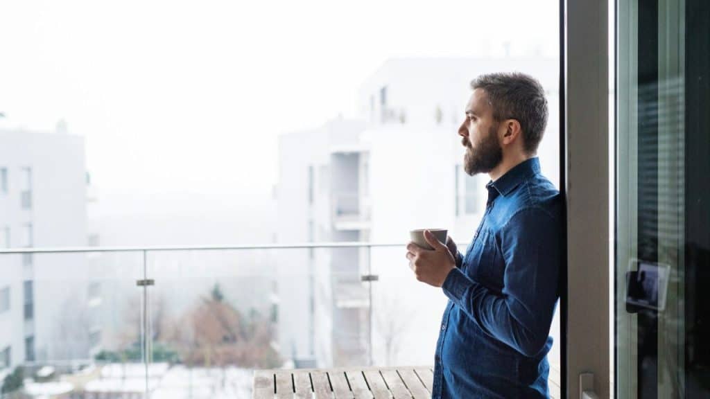 A bearded man stands on a balcony holding a mug, looking out at modern buildings.