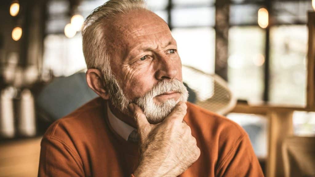 Older man with white beard and orange sweater, looking thoughtful with his hand on his chin.