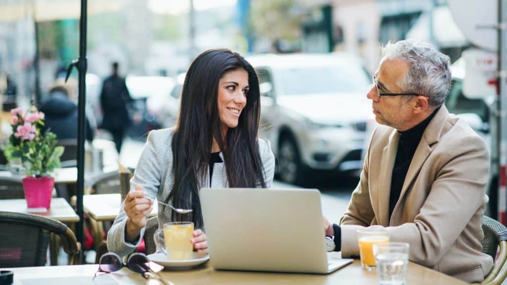 Woman smiles at man while sitting at a table with a laptop and drinks outdoors.