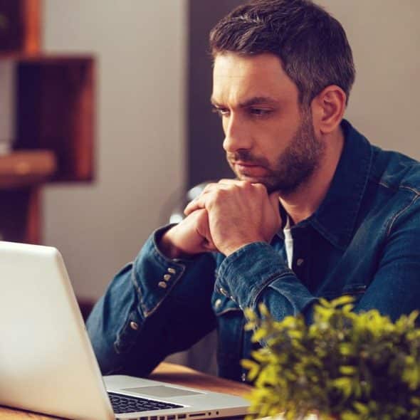 Middle-aged man in a denim shirt intently looking at a laptop screen indoors.