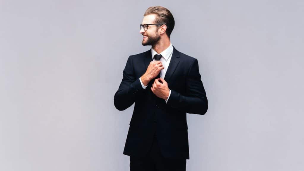 Young man in a black suit and glasses smiles while adjusting his tie.