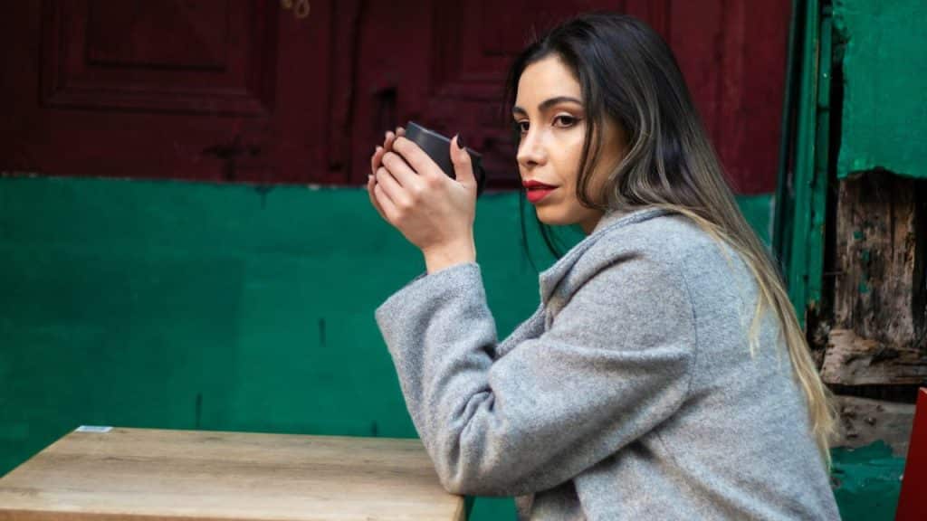 A woman in a grey coat sits at a wooden table, holding a black cup and looking off to the side.