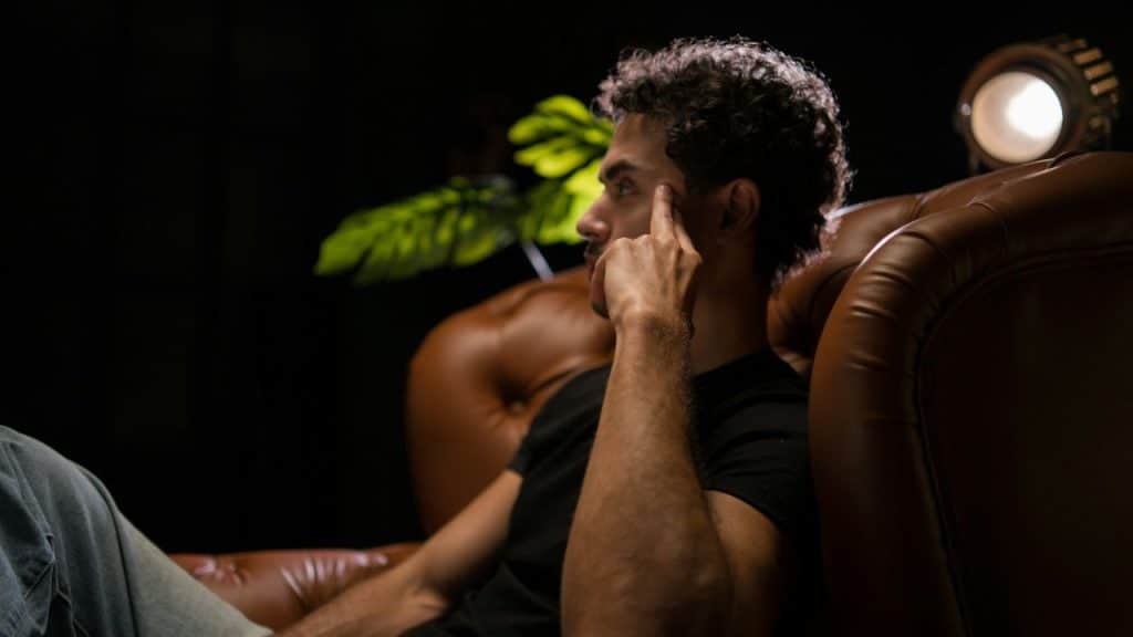 A man with curly hair sits in a leather armchair in a dark room, touching his temple with his finger.