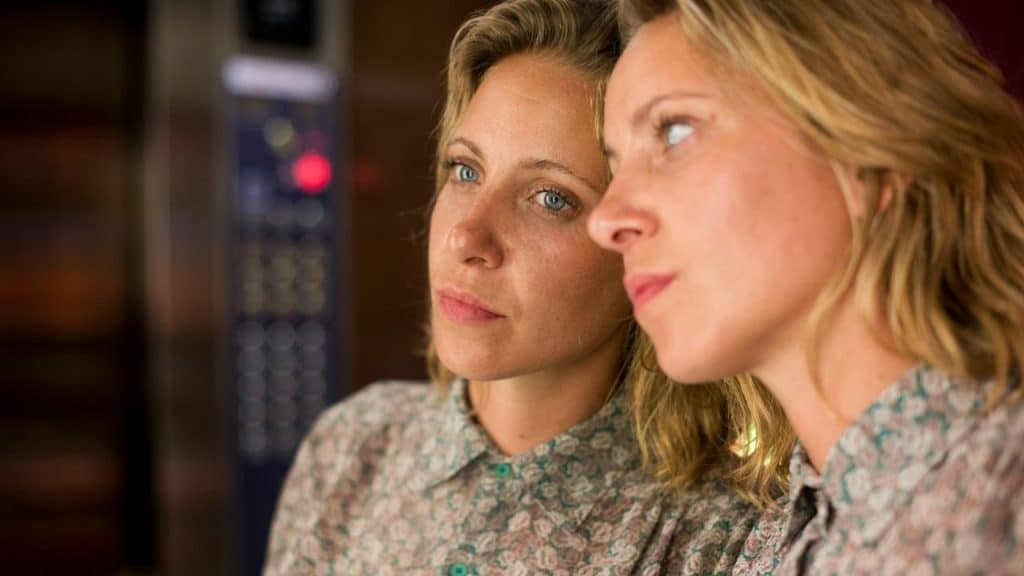 A blonde woman in a floral shirt is reflected in a mirror or glass, looking away with a pensive expression.