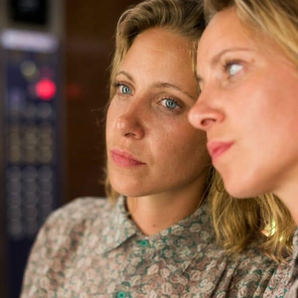 A blonde woman in a floral shirt is reflected in a mirror or glass, looking away with a pensive expression.