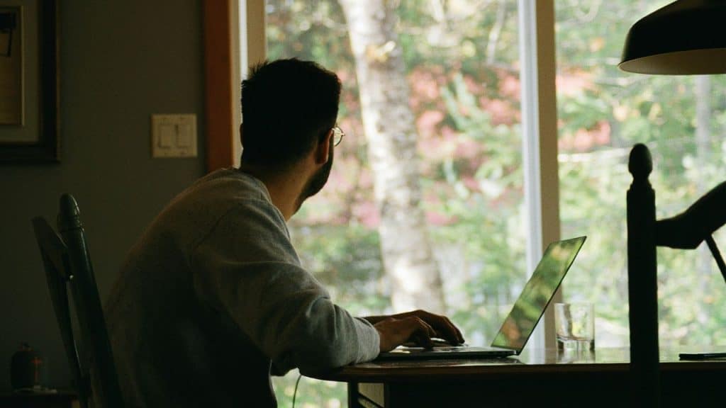 A man in glasses and a grey sweater is sitting at a table, facing away from the camera, looking out a large window while typing on a laptop.