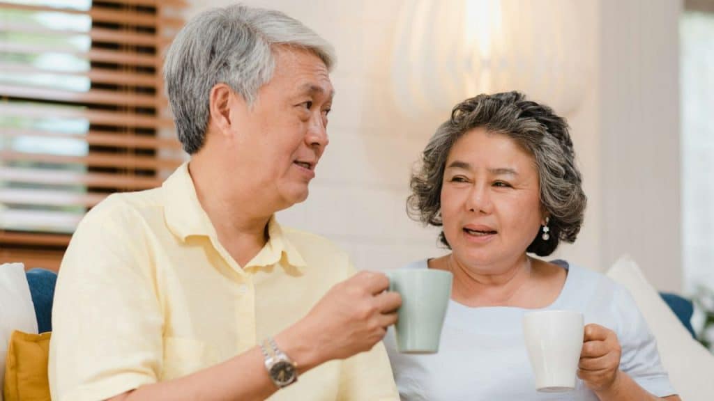 A middle-aged or elderly Asian couple holding mugs and talking in a living room.
