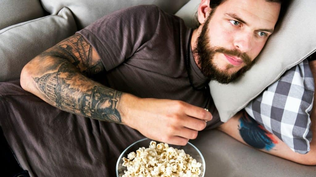 Bearded man with tattoos lying on a couch eating popcorn from a bowl.