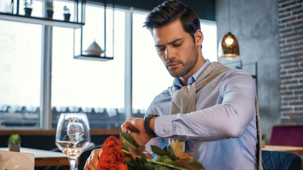 A handsome man in a blue shirt and a sweater draped over his shoulders looks at his watch while seated at a table with roses.