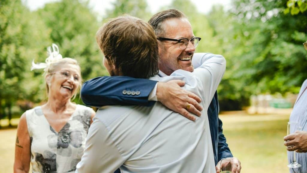 Two men hugging outdoors; a smiling woman watches in the background.
