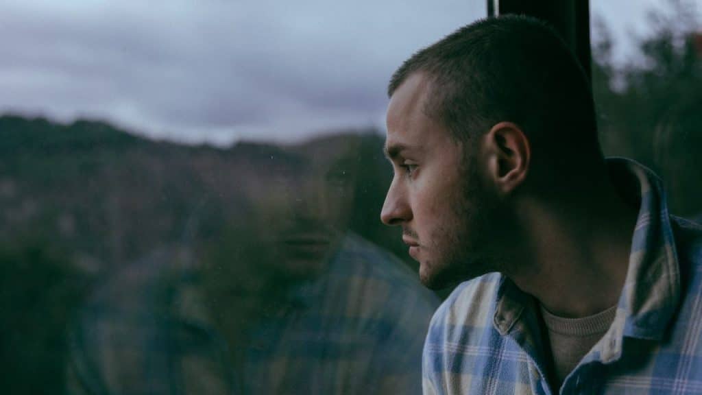 Man in a plaid shirt looking out a window with his reflection visible.
