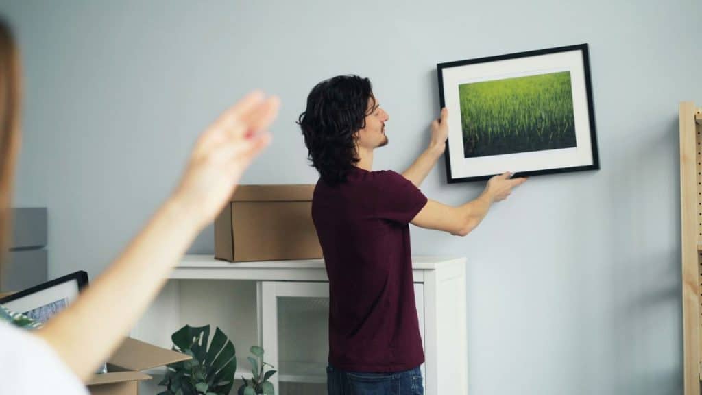 A man with long dark hair is hanging a framed picture on a wall while a person with a raised hand is standing in the blurry foreground.
