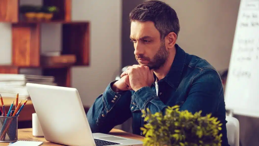 A serious man in a denim shirt is leaning on his hands and looking down at a laptop.