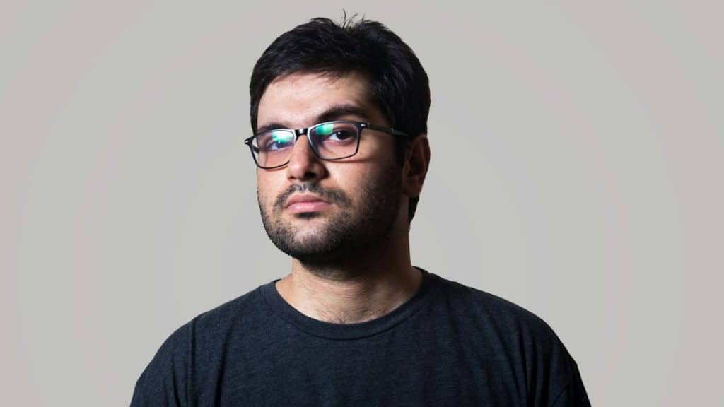 Young man with dark hair, beard, and glasses looks directly at the camera.