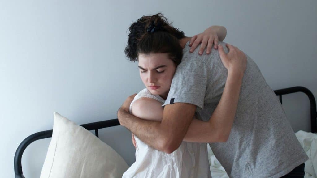 Two people embrace on a bed; one looks down with a distressed expression.