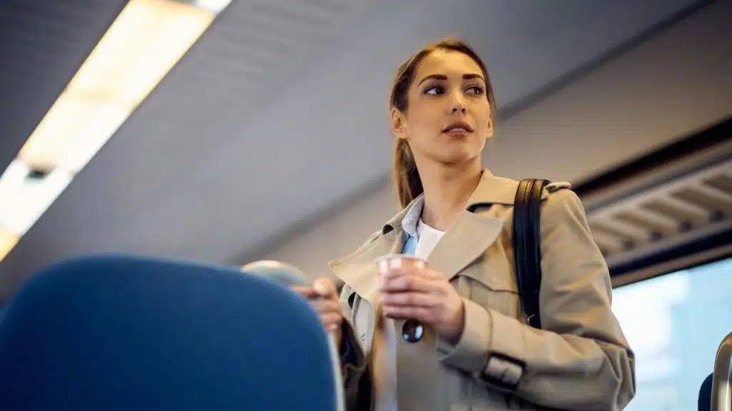 Woman in a trench coat, holding a cup, stands inside a train or subway car.