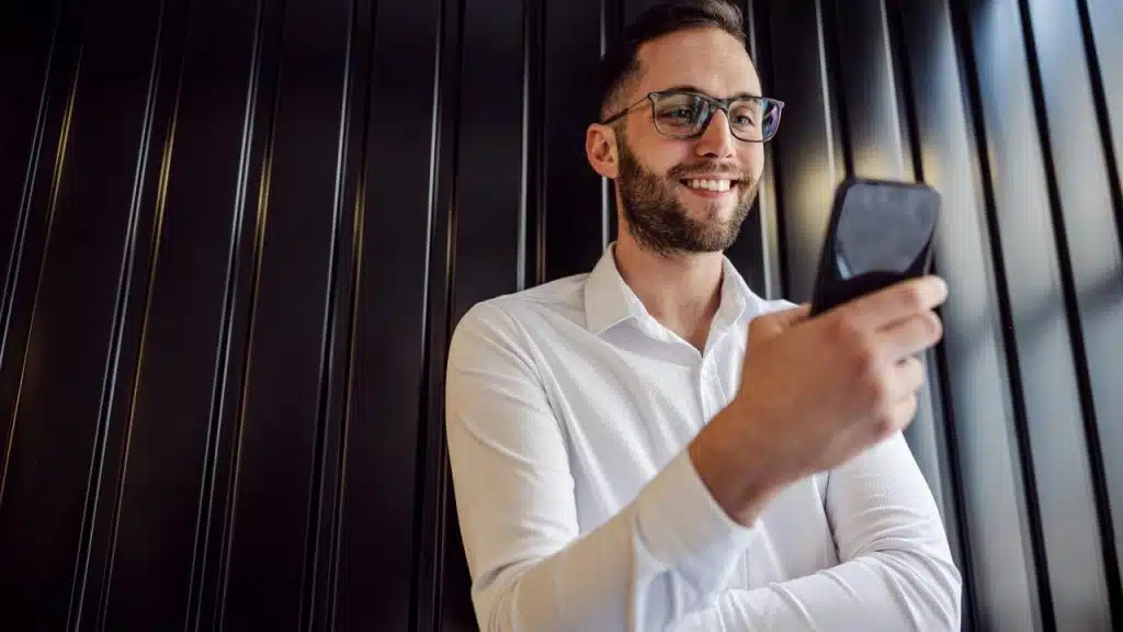 A man wearing glasses and a white shirt smiling while looking at his phone.