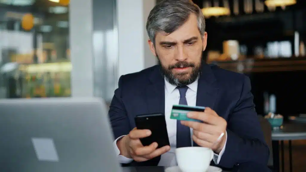 A man in a suit is seated at a table, holding a phone and a credit card near a laptop and coffee cup.