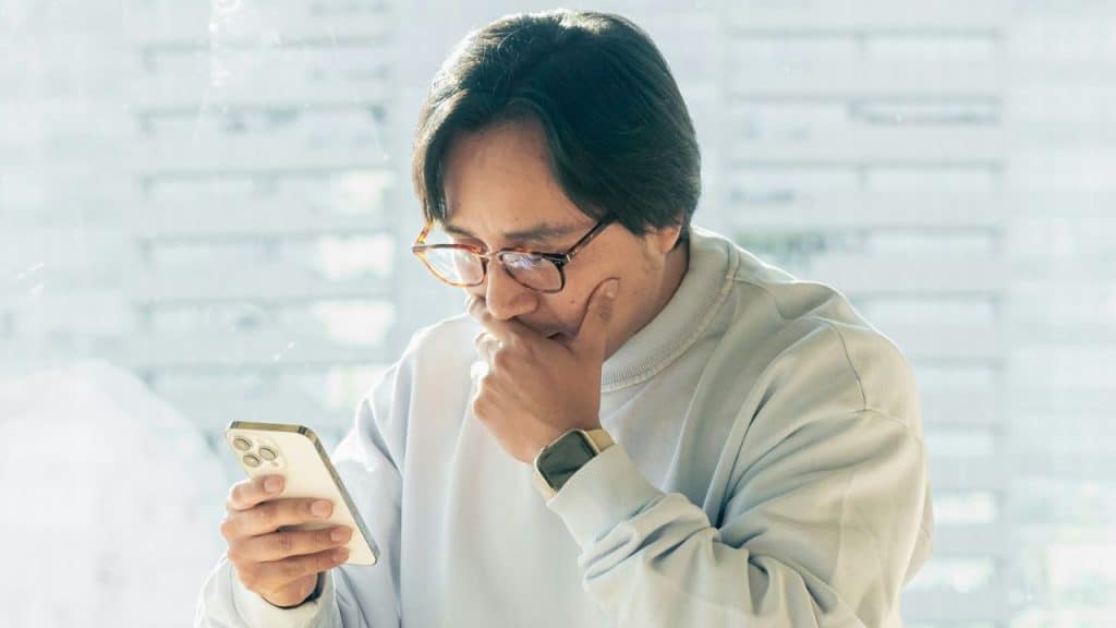 Man with glasses looking intently at a smartphone, hand near his chin.