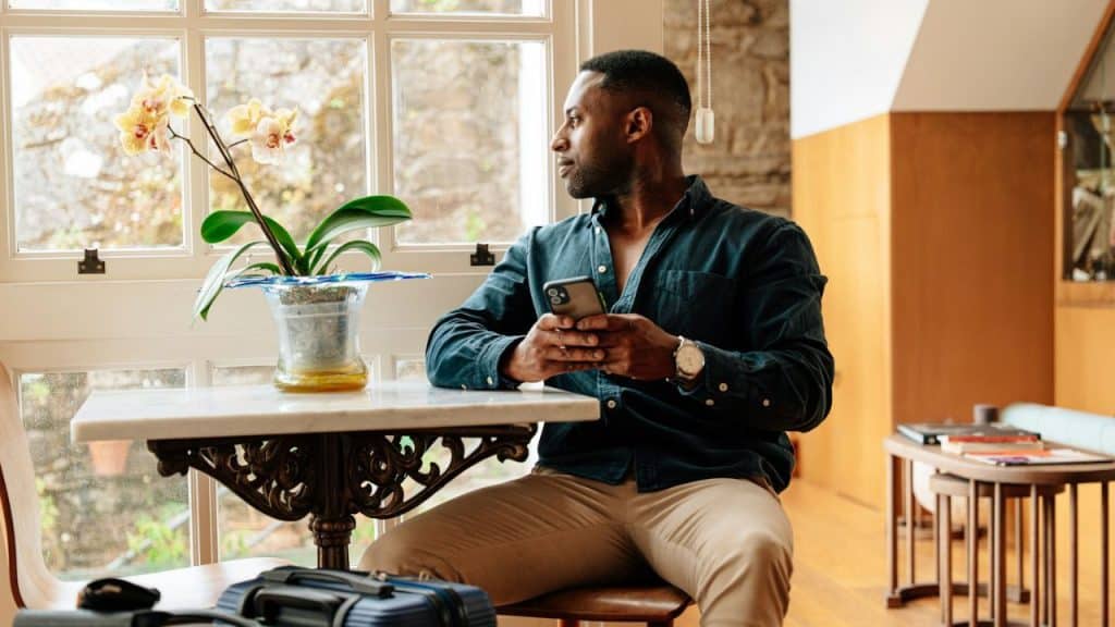 Black man with phone looks out a window next to an orchid plant on a table.