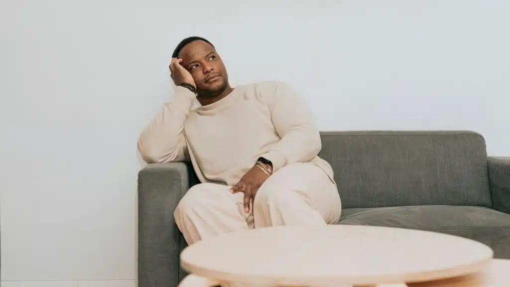 Black man in beige loungewear sitting on a couch with his head in his hand.