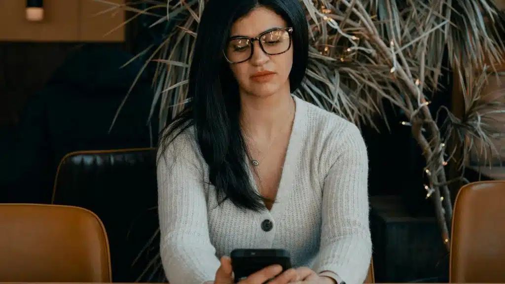A woman with dark hair and glasses looking at her phone in a café.