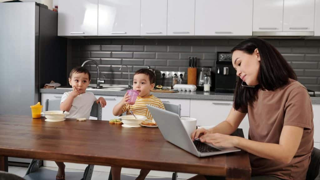 A mother working on a laptop while her children eat breakfast.