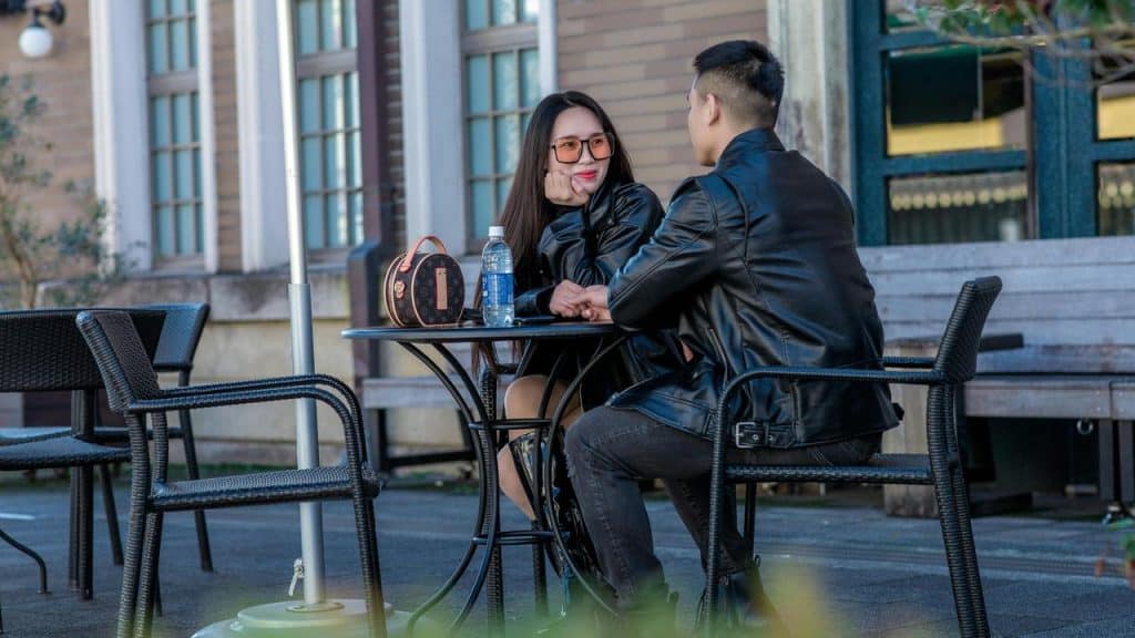 A couple sitting at an outdoor café table holding hands and smiling at each other.