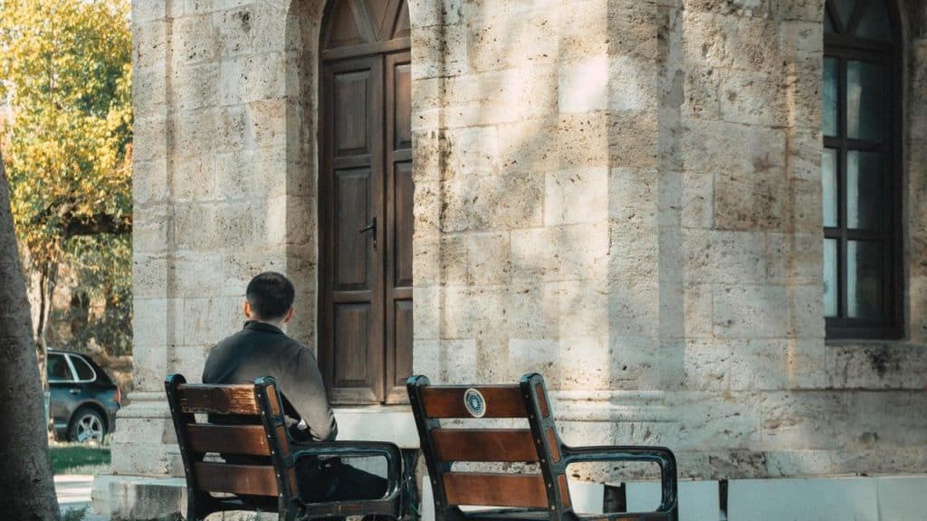 A person sitting alone on a bench outside a stone building with wooden doors.