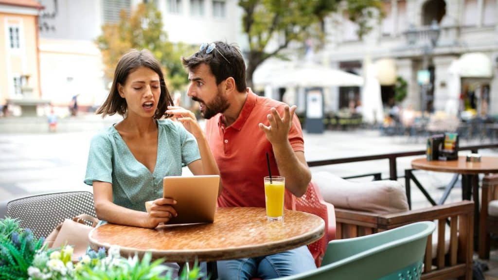 A frustrated couple argues at an outdoor café table, where the woman is holding a tablet.
