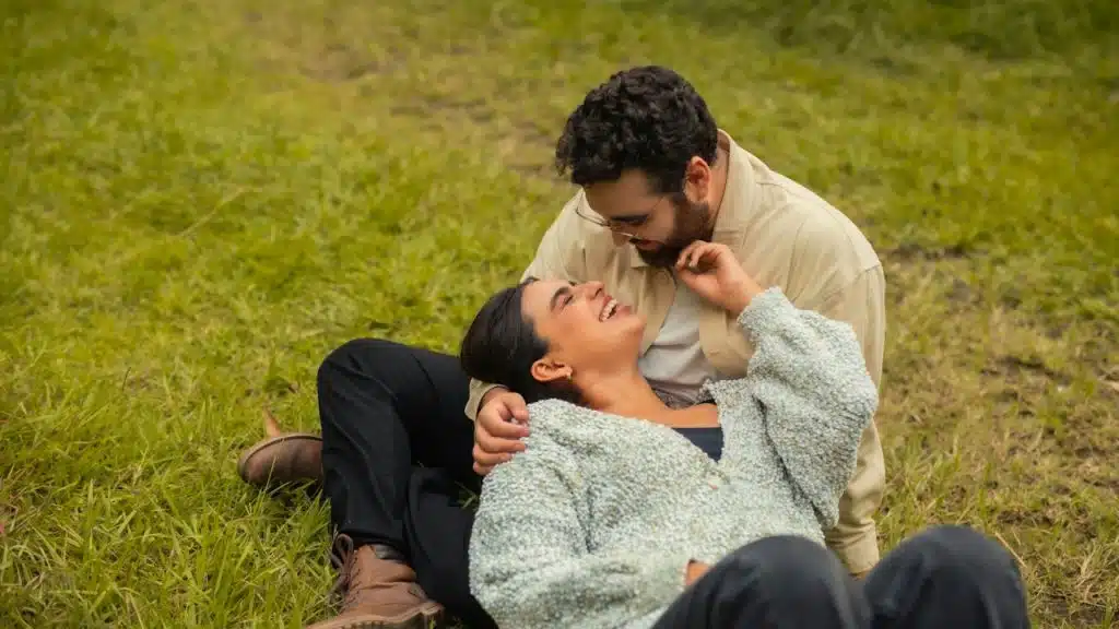 A happy couple outdoors, with the woman lying back on the grass and laughing, and the man leaning over her, smiling while touching her face.