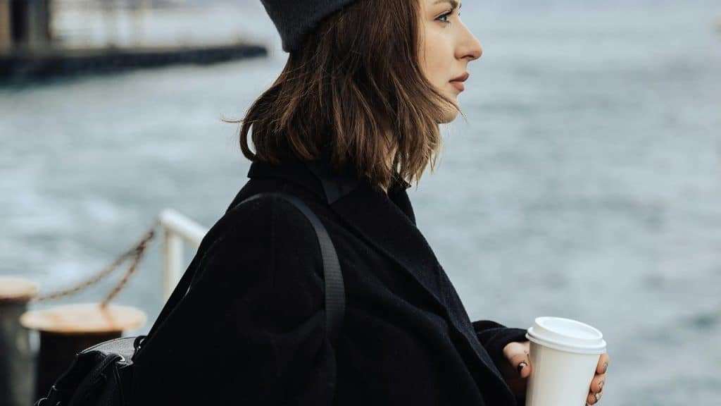 A woman in a coat and beanie holding a coffee cup by the water.